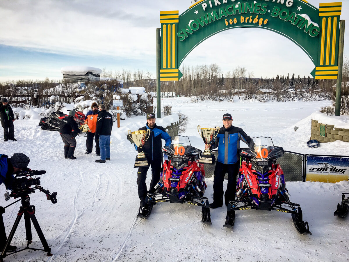 Two men in heavy winter gear stand next to snowmachines (snowmobiles) near a green-and-yellow arch that marks the finish line of the Iron Dog Snowmachine Race.