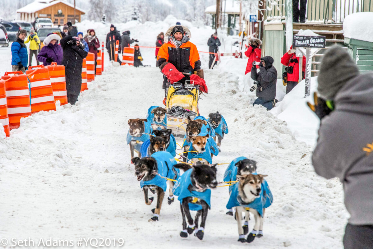 Sled dog team and musher in orange parka run down snowy chute.