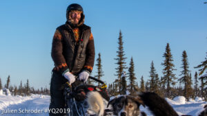 Smiling man wearing winter clothes stands atop a sled passing by, driven by sled dogs along a snowy forest trail.