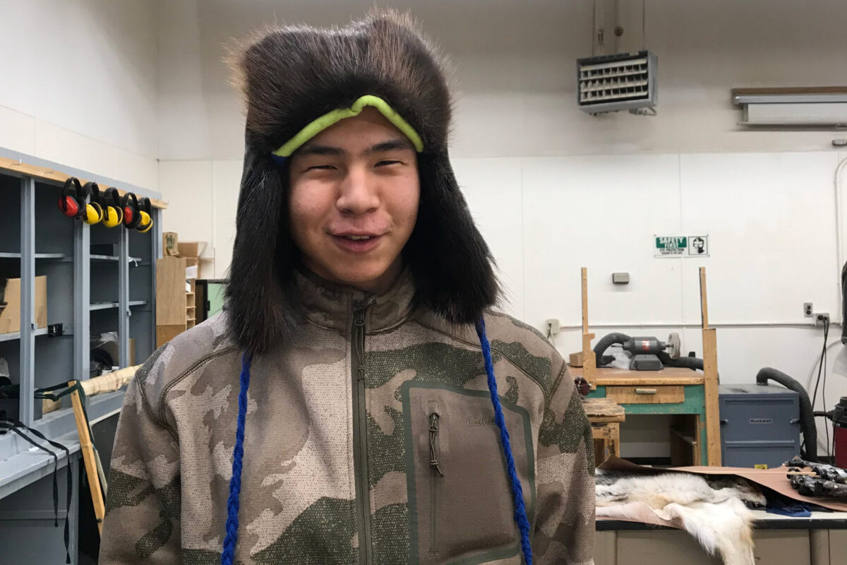 Teenaged boy, wearing a camouflage jacket, poses in high school workshop wearing handmade, fur winter hat.