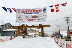 Looking down a snow-covered street in a rural Alaska town with a large "Iditarod" banner hanging overhead and a carved wooden archway standing in the center of the street.