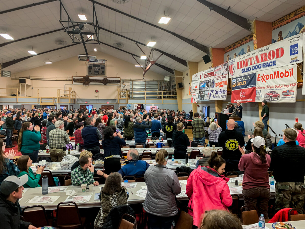 Iditarod fans stand and cheer Pete Kaiser in a large gymnasium, converted into a banquet hall.