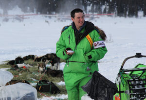 Man in bright green parka stands in front of a sled dog team resting on the ground.