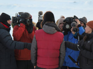 Musher in red vest, seen from behind, answers questions to a large group of closely-gathered reporters, many holding cameras or microphones.