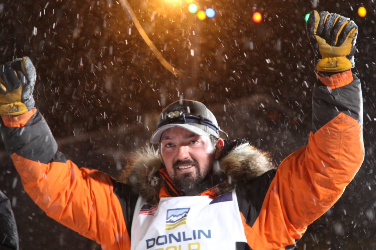 Man in orange and black parka raises his hands in triumph after his Iditarod victory.