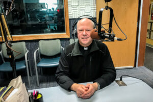 Bishop smiles at camera while seated inside radio studio behind microphone.