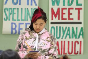 a young girl writes on a notepad with a pencil, with posters behind her that read "Spelling Bee" and "Quyana"
