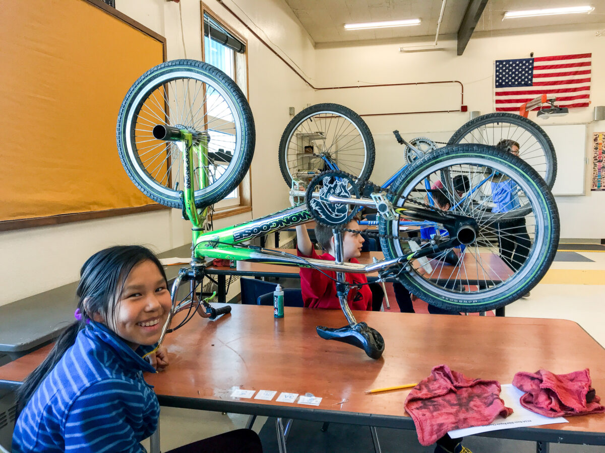 A student smiles at the camera, seated at a table on which stands a bicycle being repaired.