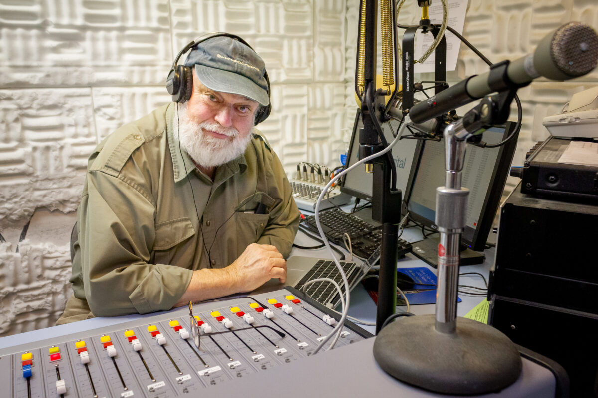 Man with white beard sits in front of sound board inside radio broadcast studio