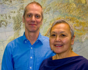 Man and woman stand smiling in front of a large wall map of northwestern Alaska.