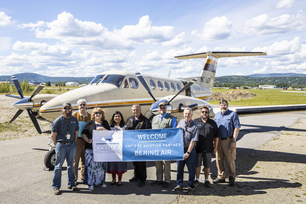 Smiling group of people stands in front of a small airplane on a runway on a sunny, summer day