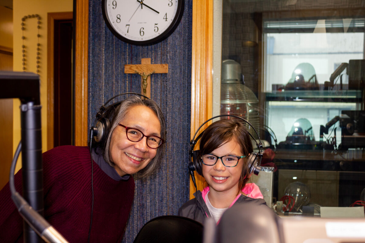Middle-aged woman and young child smile from behind radio microphone inside studio