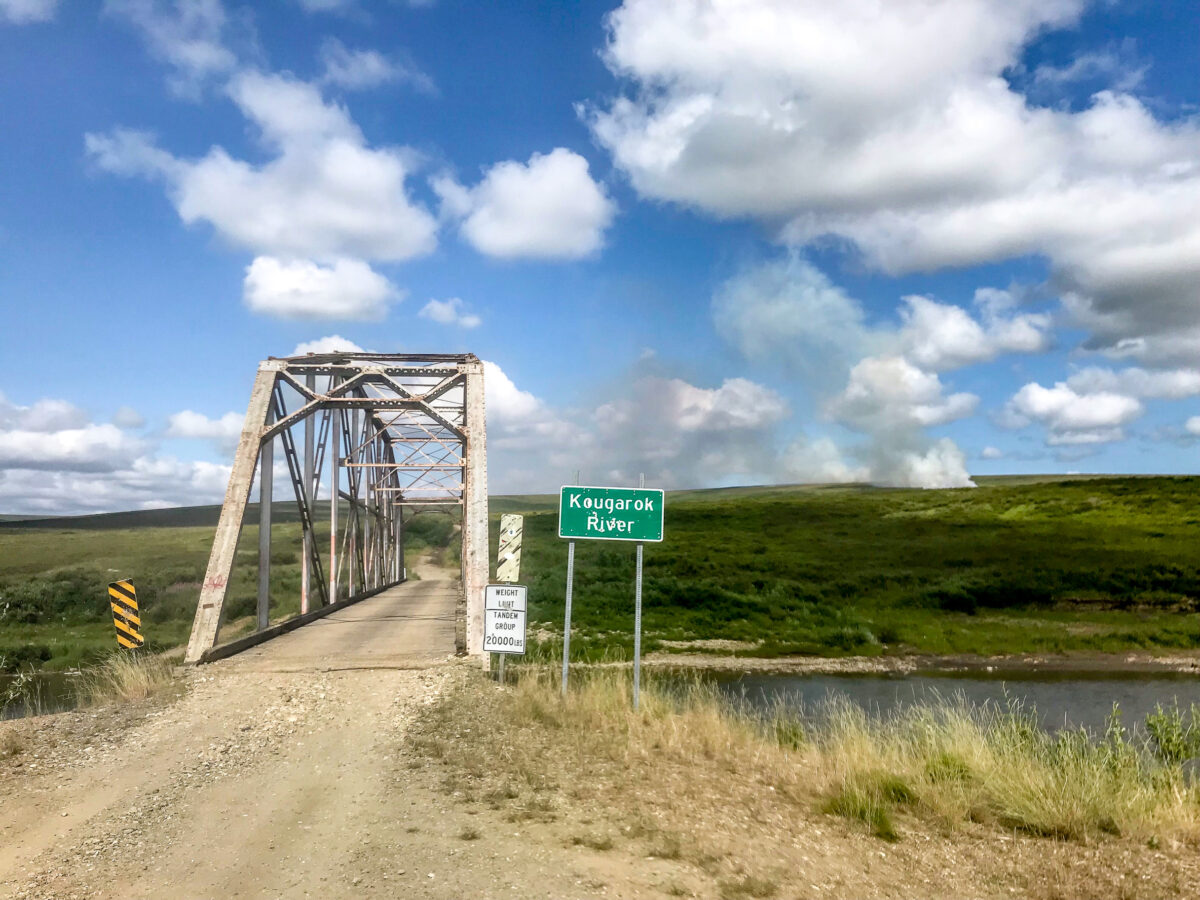 A landscape of a metal bridge, creek, and tundra stretching to the horizon, with puffs of smoke arising from a wildfire about a half-mile away.