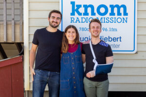 Three young people stand smiling in front of a sign that reads "KNOM Radio Mission."