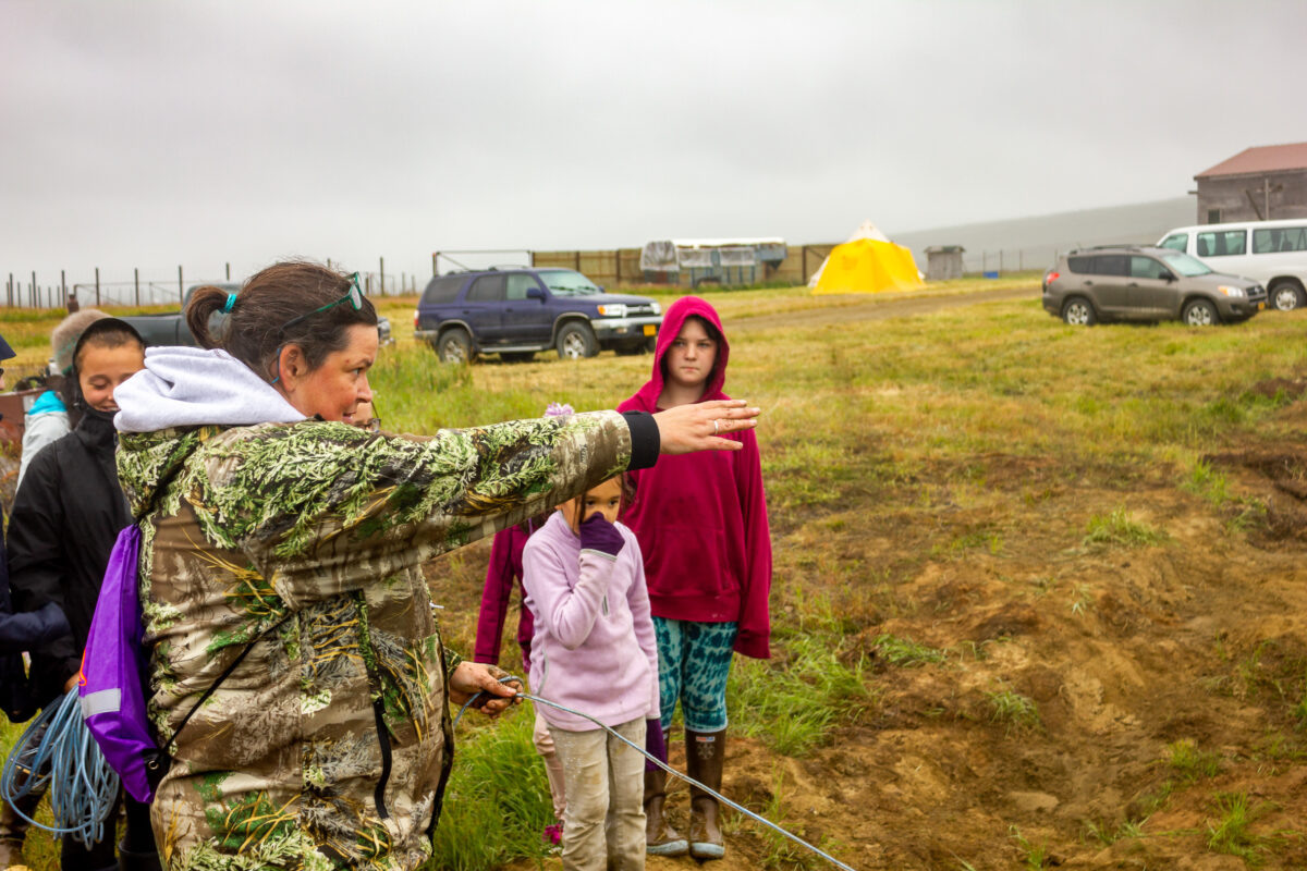 Woman in camouflage jacket stands on grassy tundra next to huddle of children.