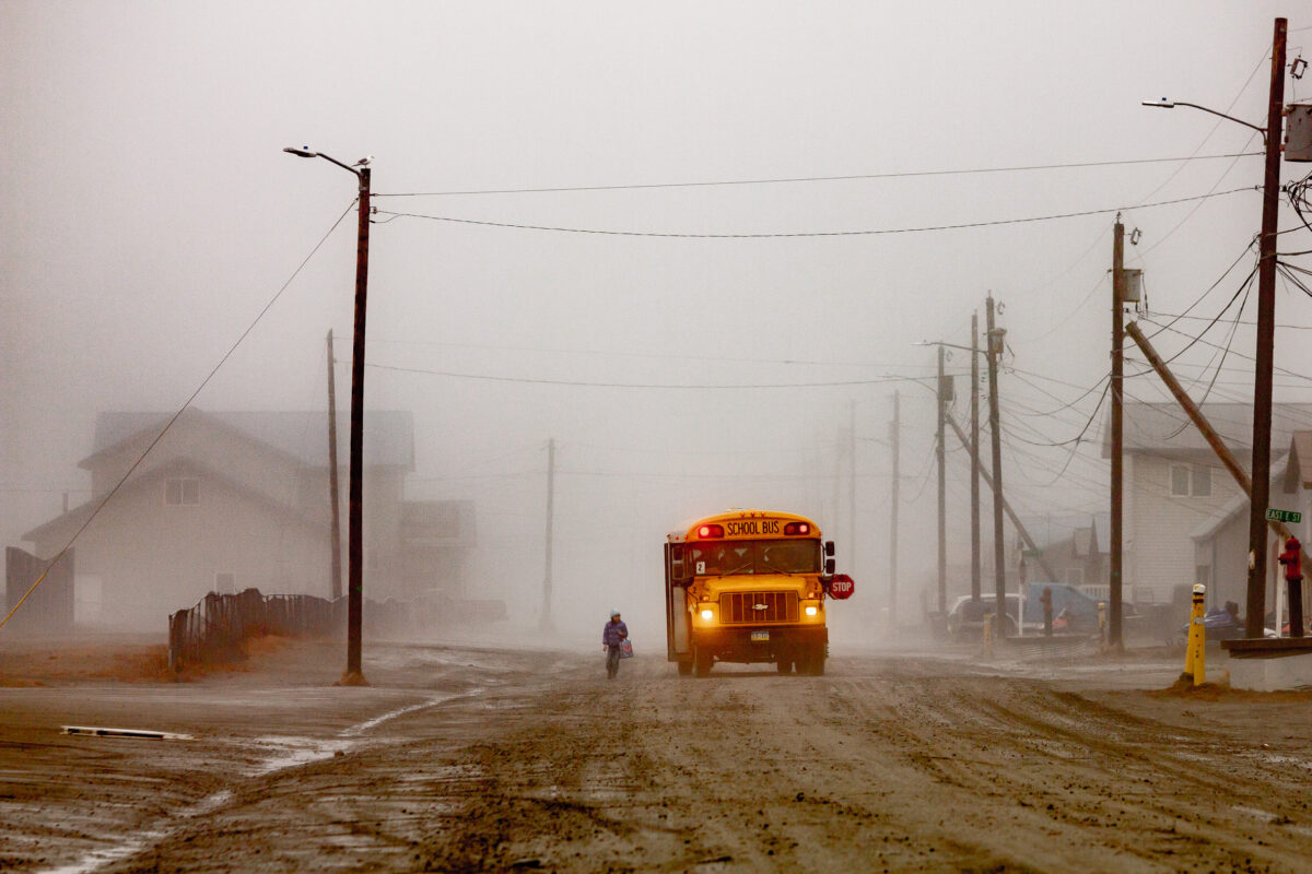 Amid heavy fog, a yellow school bus is stopped in the middle of a dirt road in a rural Alaska town, waiting for a young student to board.