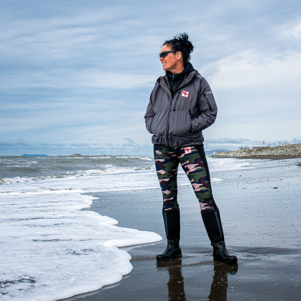 Woman in technical clothes stands on rural Alaska beach on a cloudy day