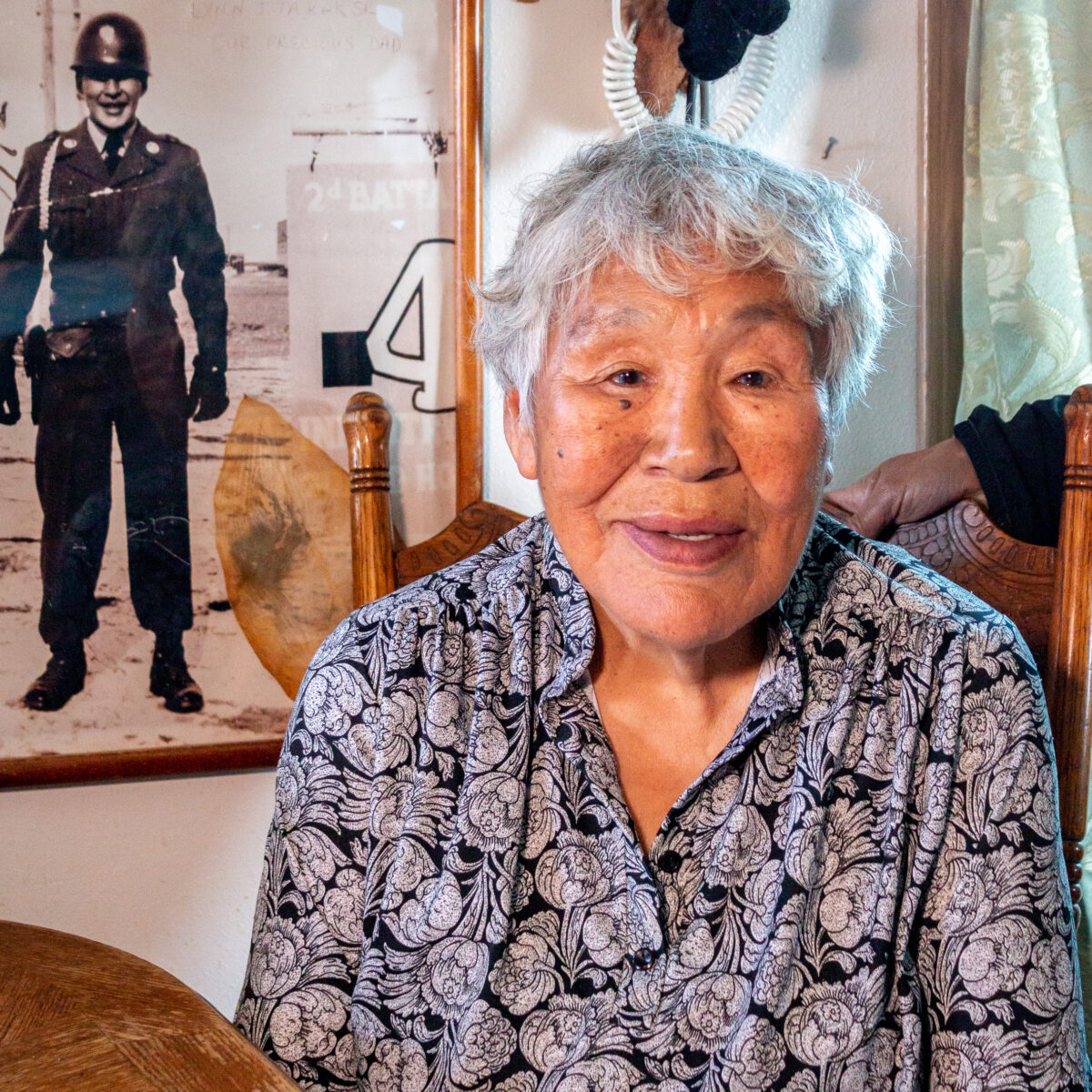 Elderly woman wearing floral-print shirt sits at table with black-and-white photo of man in uniform framed on wall behind her.