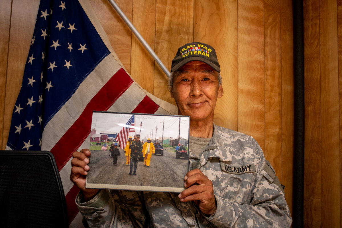 Man in Army uniform holds photo in front of American flag and wood-paneled wall