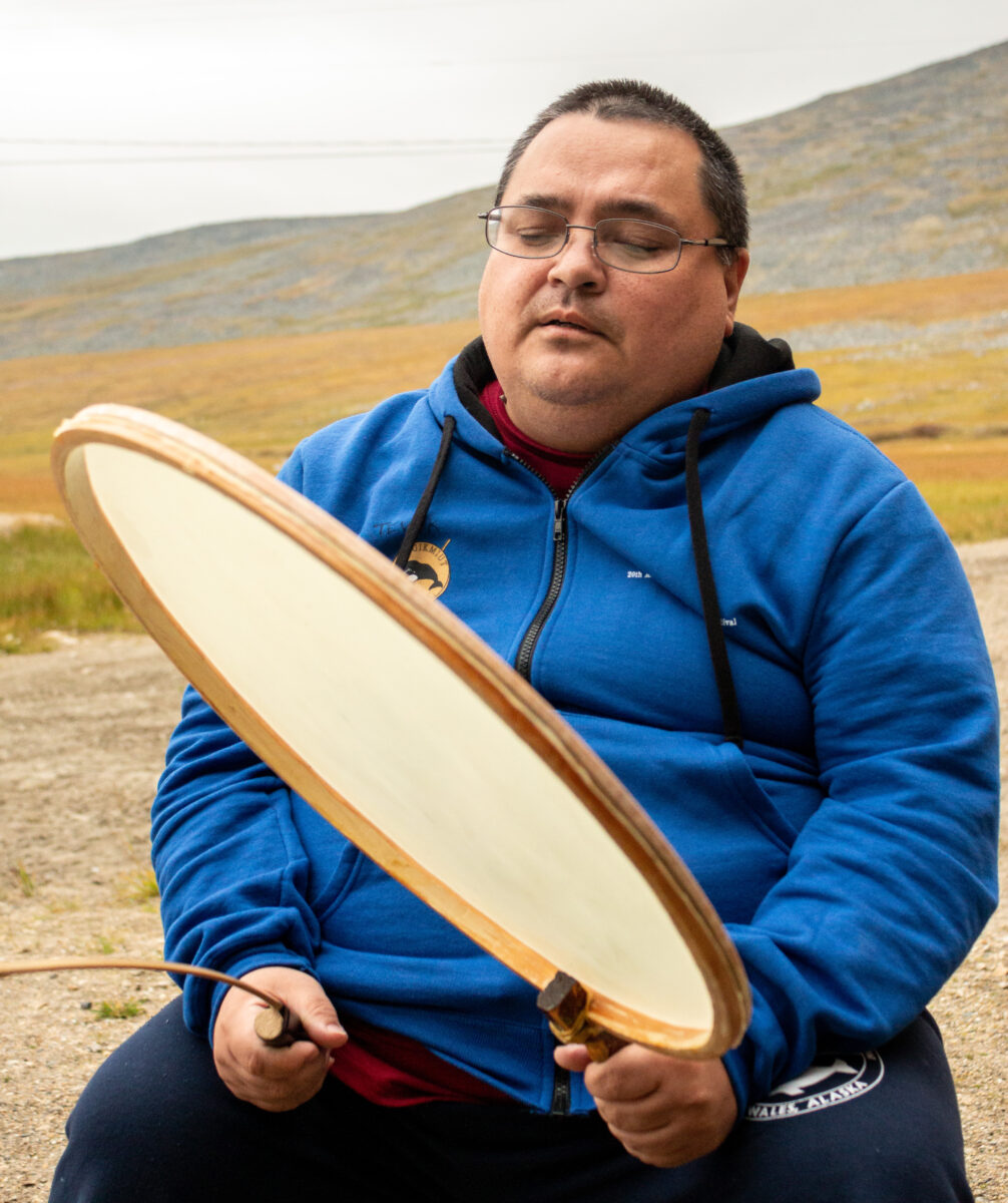 Man in blue sweatshirt performs song on traditional Alaska Native drum