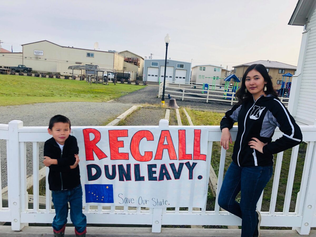 A woman and a young child stand next to a sign reading “Recall Dunleavy”