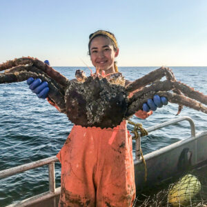 Woman in fishing gear stands on a boat at sea, holding a huge crab
