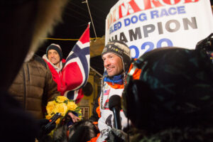 A picture of Thomas Waerner in front of the Iditarod finish sign and a Norwegian flag in Nome, Alaska.