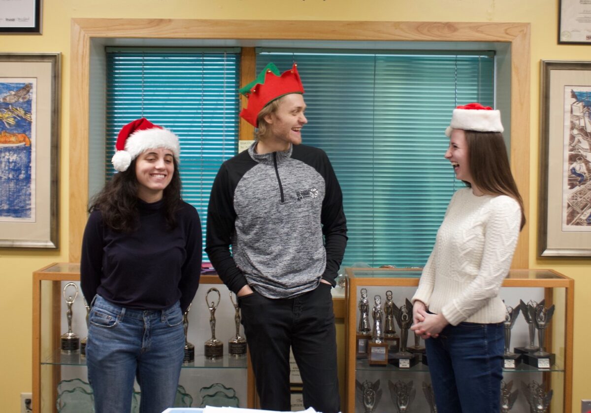 KNOM volunteers: Cathy Rubano, Colin O'Connor, and Sophia DeSalvo in the radio station wearing Christmas hats and laughing.