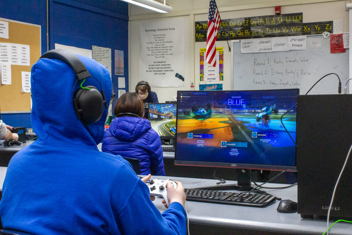 Student in blue hoody sitting at desk with a video game controller playing a game.