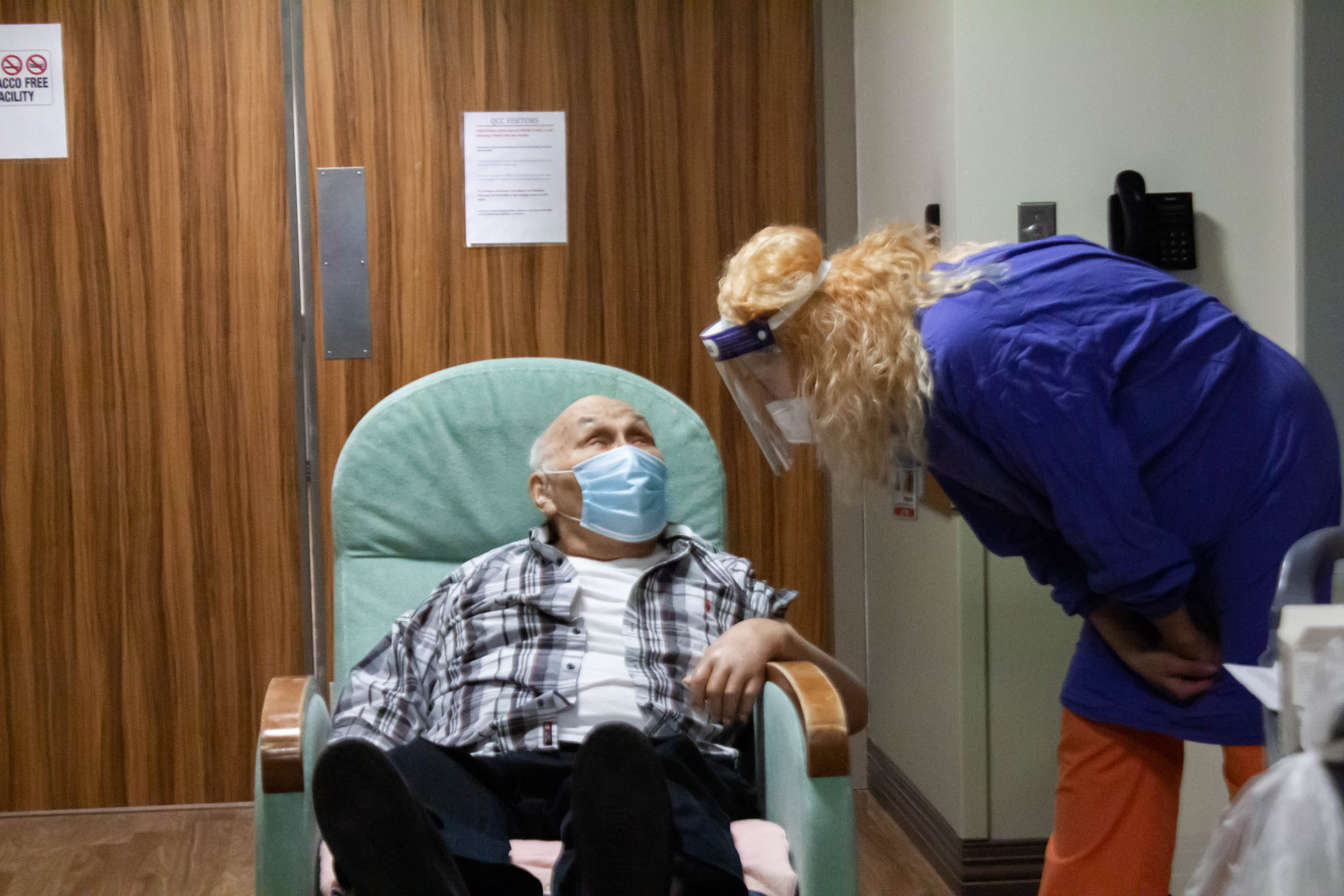 Elder man with a face mask sitting in a chair talking with nurse who is wearing a face shield