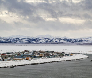 Aerial view of Unalakleet. There is mountains in the distance with water in the forefront. Buildings line the coastline. It is winter and snow is covering the ground.