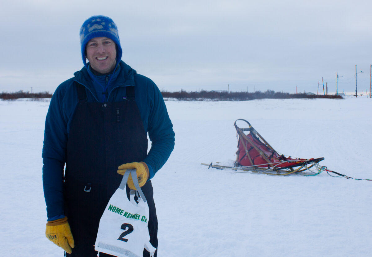 Man holding race bib in the winter time. Dog sled behind him.