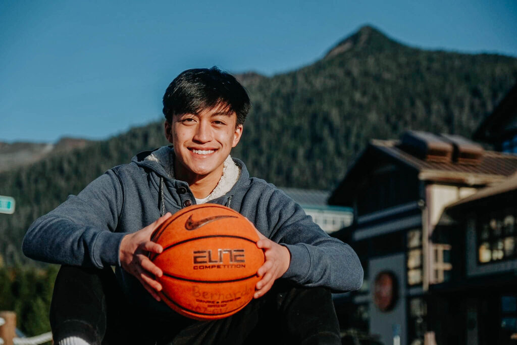 A smiling boy sitting in front with a basketball in his hands. Mountain in the background.