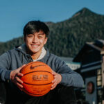 A smiling boy sitting in front with a basketball in his hands. Mountain in the background.