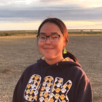 Young girl wearing glasses smiling at camera. In background, flat landscape during sunset