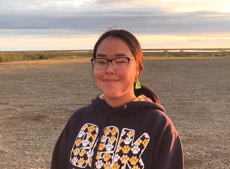 Young girl wearing glasses smiling at camera. In background, flat landscape during sunset