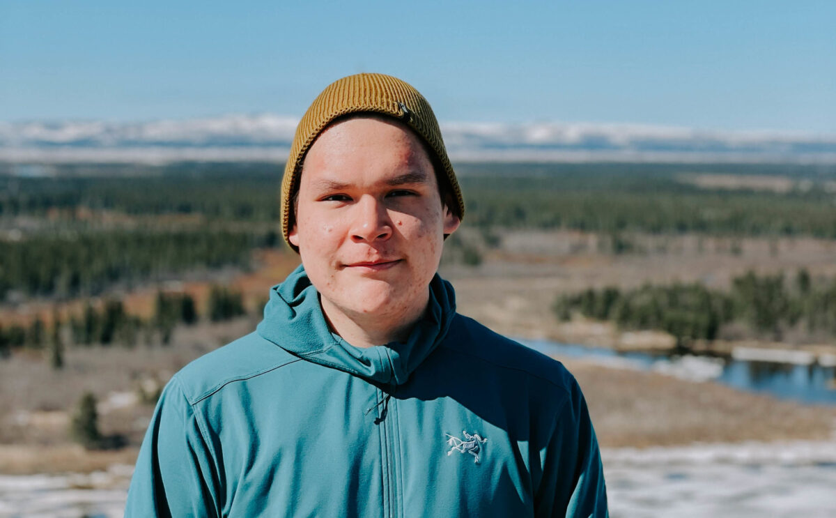 Young man looking into camera with a yellow beanie and blue jacket. Mountains and trees in the background.