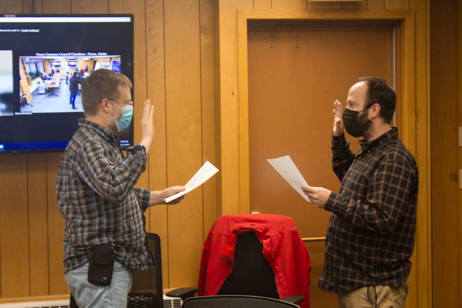 Two masked men facing each other. Both are holding a piece of paper in one hand and then have the other hand raised. They are inside of a building.