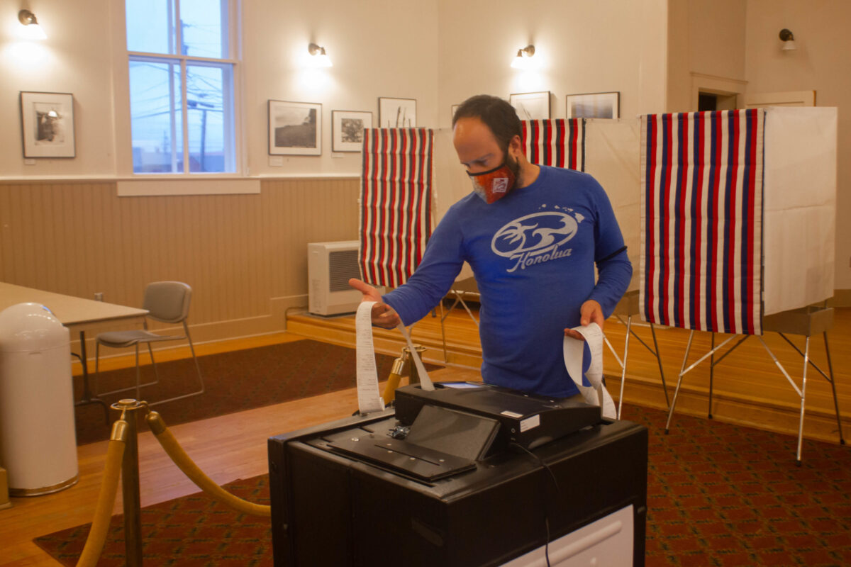 A man wearing a face mask and blue shirt holding up a long piece of paper. He is at a polling booth.
