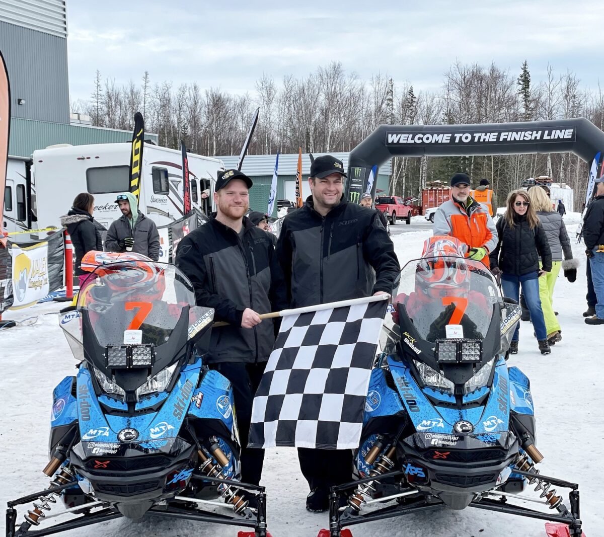 Two men standing next to the snow machines holding a checkered flag at the finish line