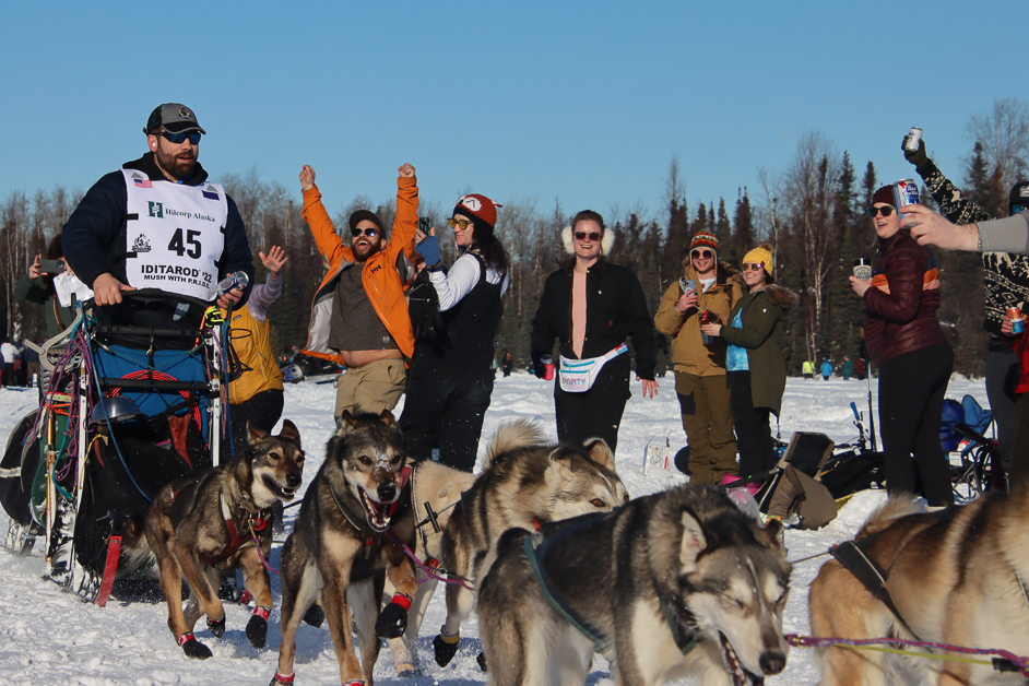 Musher racing with dogs. Crowd cheering him on.