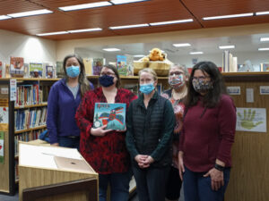 2 Women wearing mask standing for a photo in the library. One is holding a book