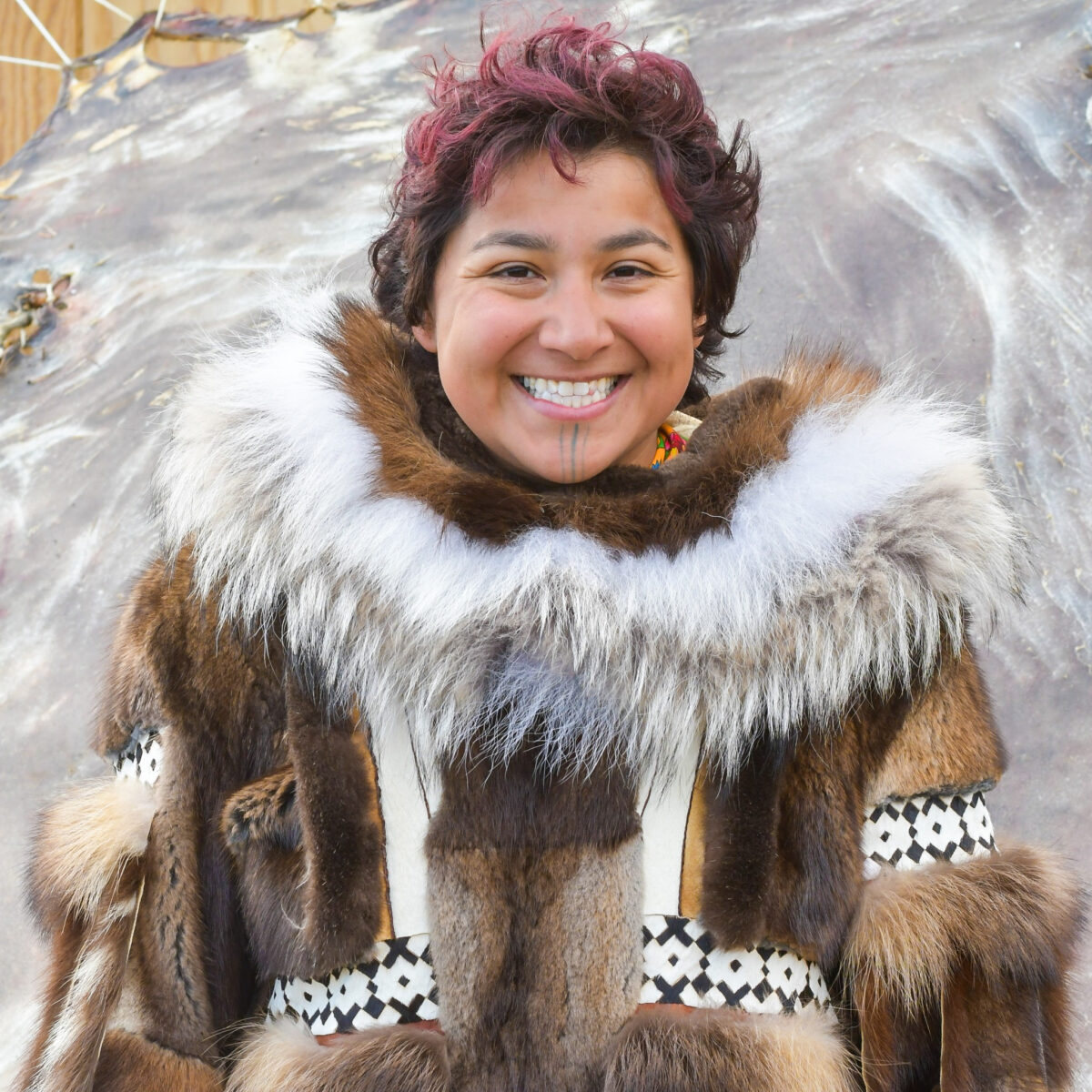 A woman, Marjorie Kunaq Tahbone, smiles, wearing a traditional Unupiaq parka. She stands in front of a stretched skin.