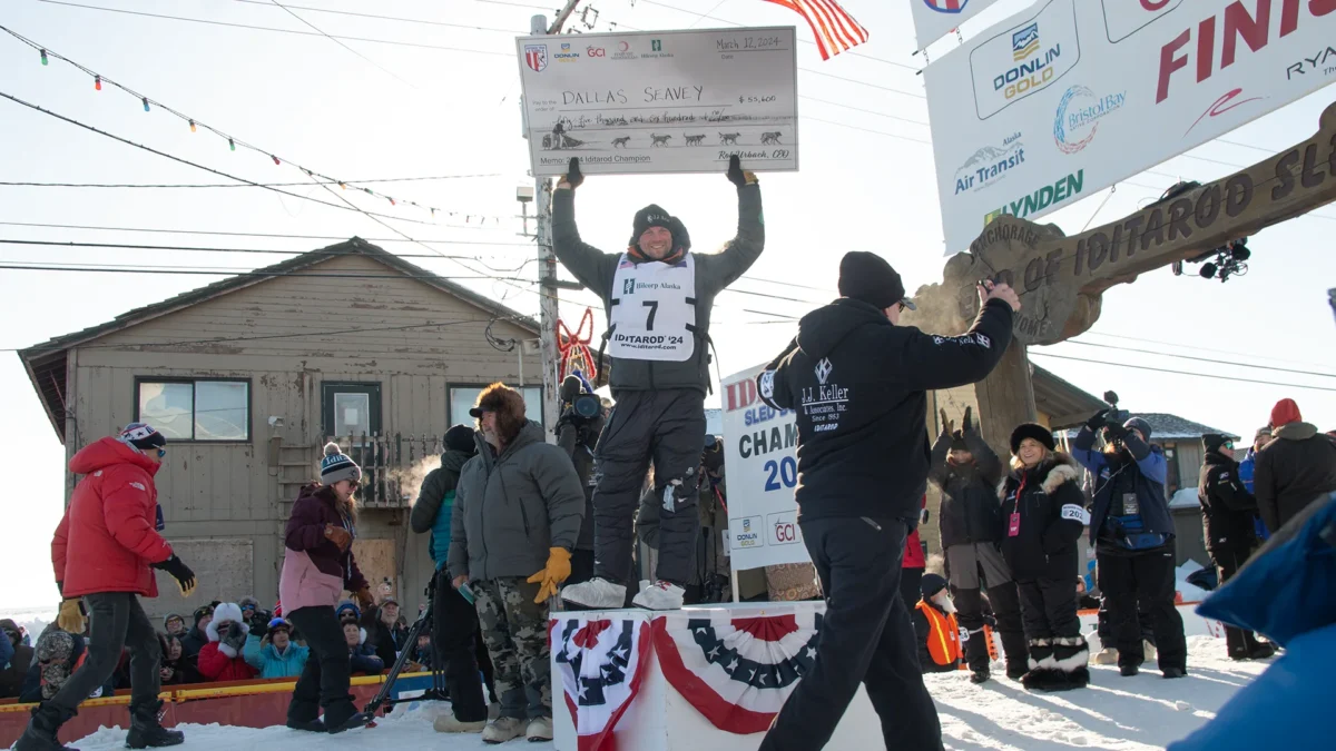 2024 Iditarod Champion, Dallas Seavey, holds an oversized check above his head as the crowd on Front Street celebrates his victory.