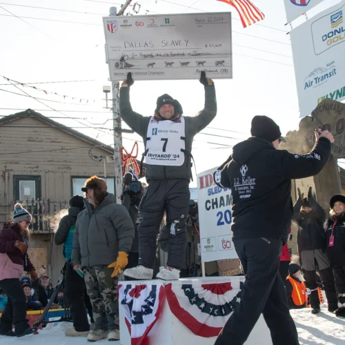 2024 Iditarod Champion, Dallas Seavey, holds an oversized check above his head as the crowd on Front Street celebrates his victory.