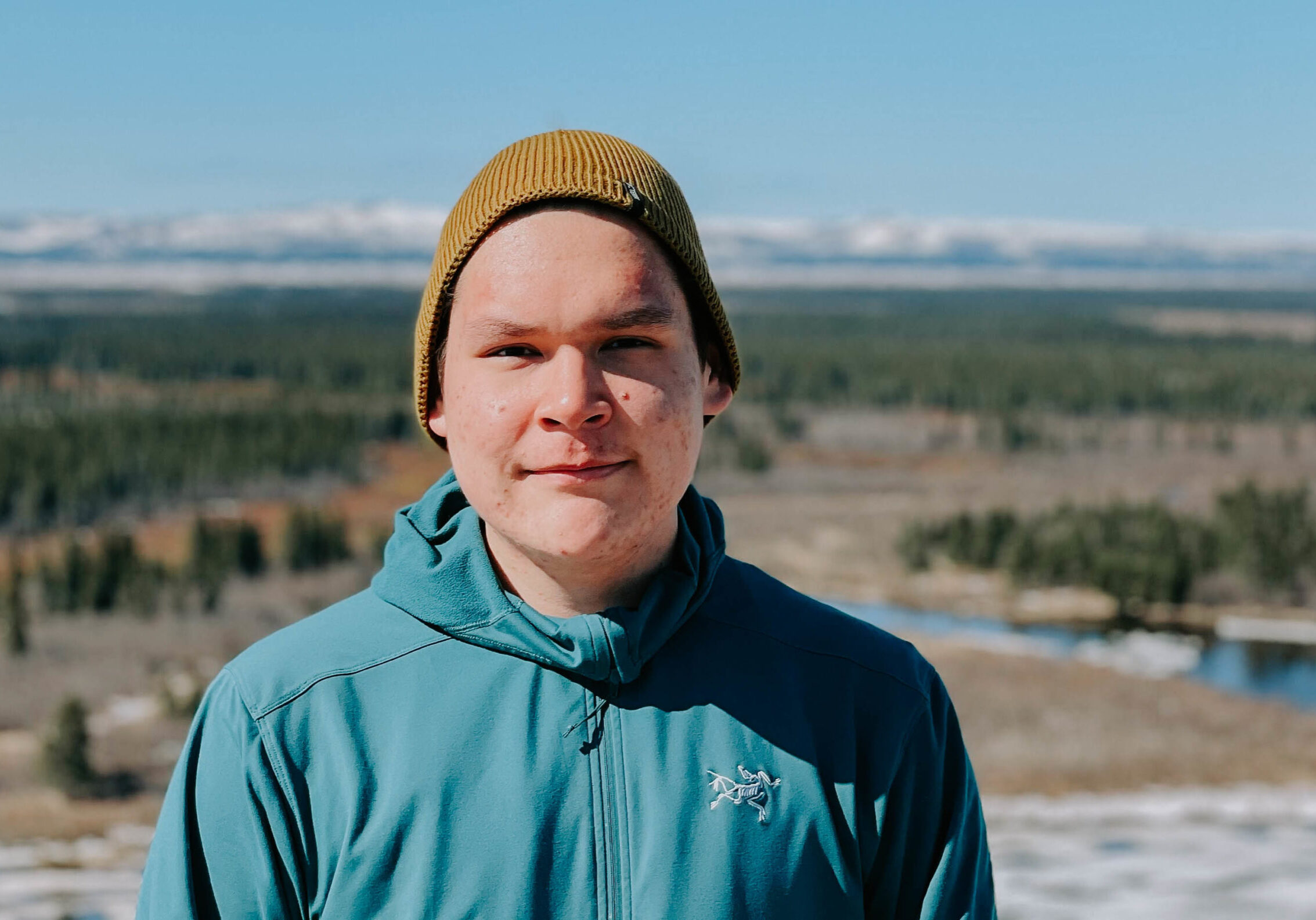 Young man looking into camera with a yellow beanie and blue jacket. Mountains and trees in the background.